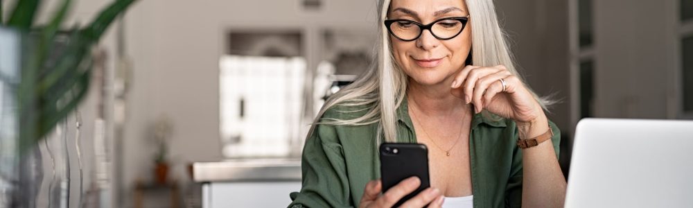 Happy senior woman using mobile phone while working at home with laptop. Smiling cool old woman with white hair wearing eyeglasses sitting on chair at table and messaging with smartphone. Beautiful stylish elderly lady browsing site on cellphone.